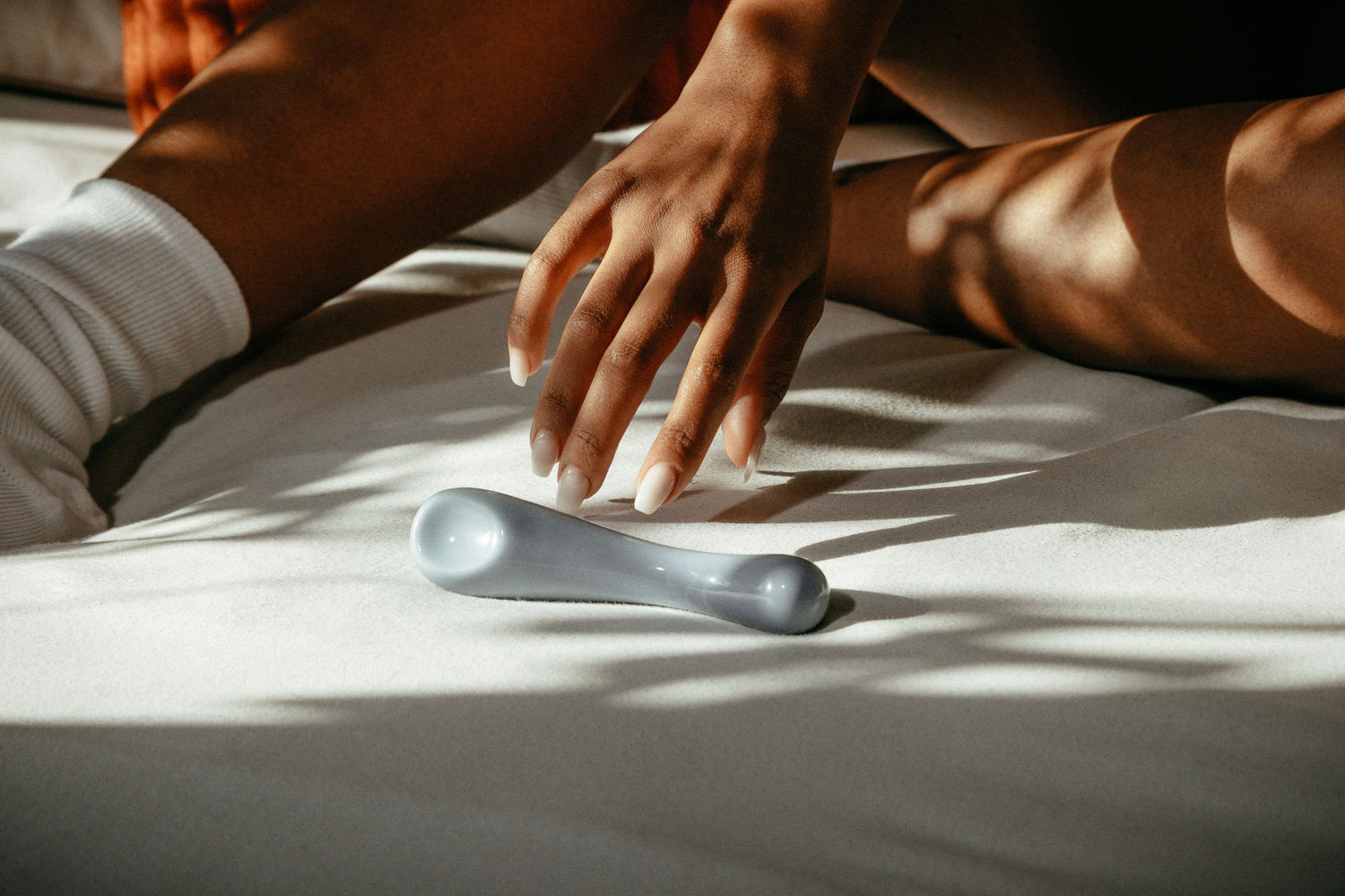 Detailbild: Zu sehen sind Körperpartien einer Person, die auf einer weichen Matratze sitzt. Im Vordergrund liegt der Dildo Clara in Blau und die Person greift mit einer Hand nach ihm