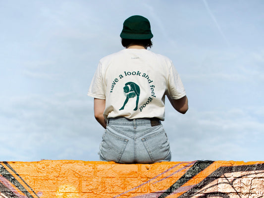 Zu sehen ist eine Person, die mit dem Rücken zum Betrachter auf einer Mauer sitzt. Sie trägt das Merchandise T-Shirt vom Studio Labim