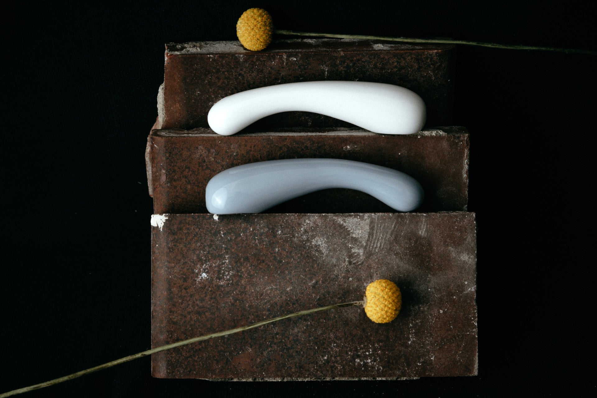 Studio Labim: Abgebildet sind die Dildos "Clara" aus Porzellan in den Farben weiß und blau. Die Sextoys ruhen für das Foto auf Ziegelsteinen auf zwei Ebenen und sind mit gelben Blumen dekoriert. 