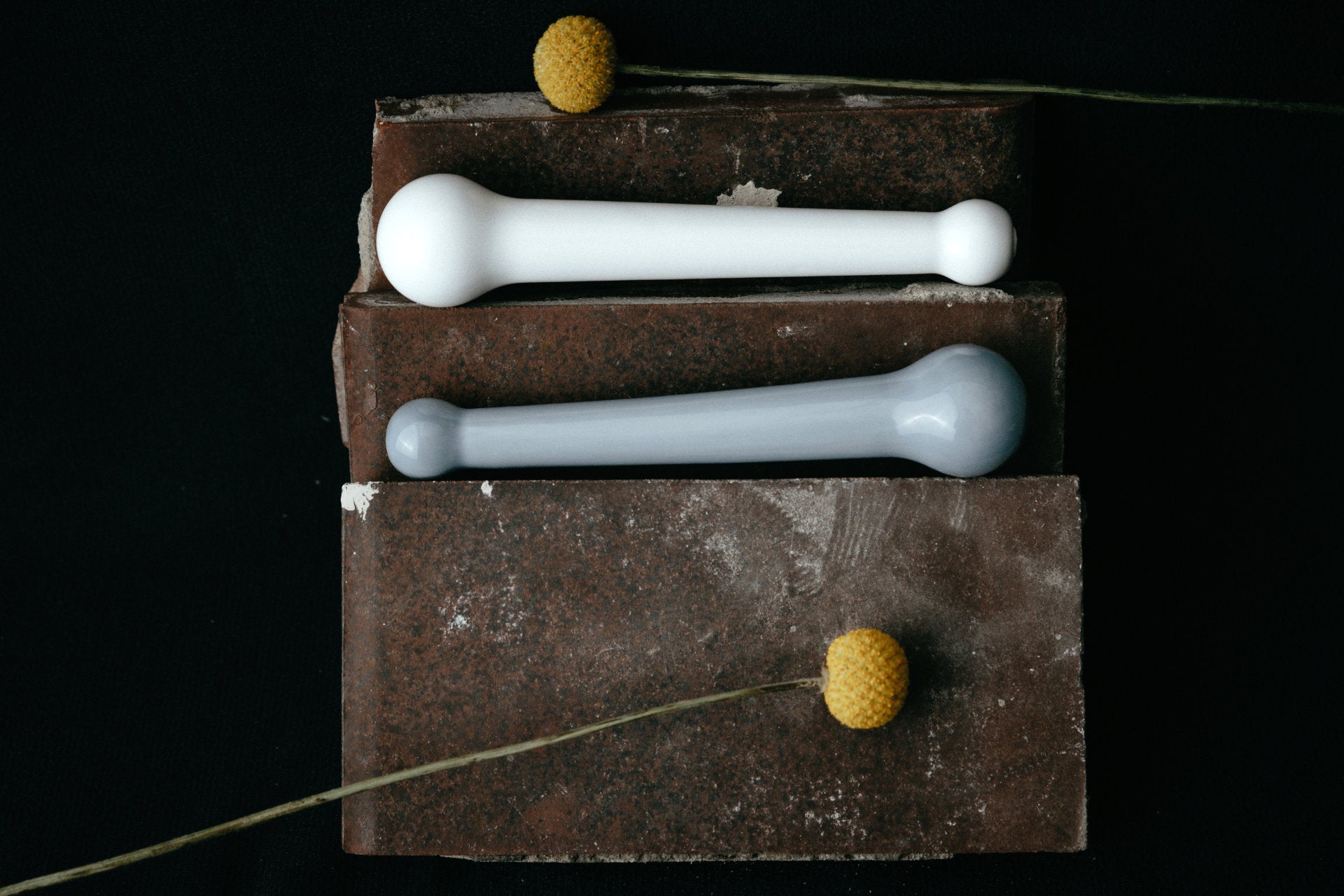 Studio Labim: Abgebildet sind die Dildos "Helen" aus Porzellan in den Farben weiß und blau. Die Sextoys ruhen für das Foto auf Ziegelsteinen auf zwei Ebenen und sind mit gelben Blumen dekoriert. 